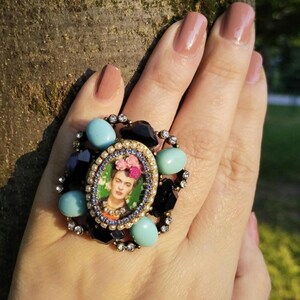 Peut inclure: Une bague imposante ornée d'un portrait de Frida Kahlo. La bague est ornée de pierres turquoise et noires, ainsi que de petits cristaux transparents. La bague est montée sur une base en métal doré, créant un accessoire audacieux.