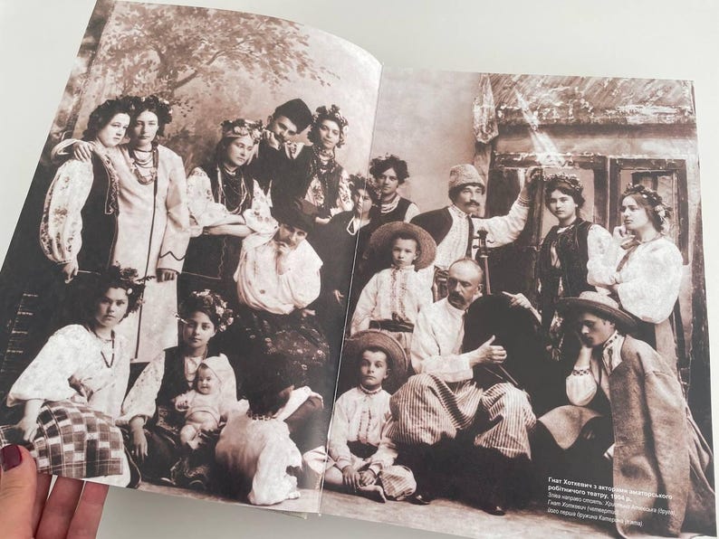 May include: A black and white vintage photograph of a large group of people, mostly women and children, standing in front of a building. The people are dressed in traditional clothing and some are holding flowers. The text "Grest Kholmova and a group of musicians performing folk songs, 1934.  A group of musicians from the village of Kholmovo, near the city of Orel, Russia.  The musicians are playing traditional Russian instruments, such as the balalaika, the domra, and the accordion." is printed at the bottom of the photo.