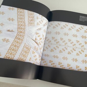 May include: Two pages from a book showing examples of white fabric with gold embroidery. The embroidery on the left page features a geometric pattern. The embroidery on the right page features a floral pattern.