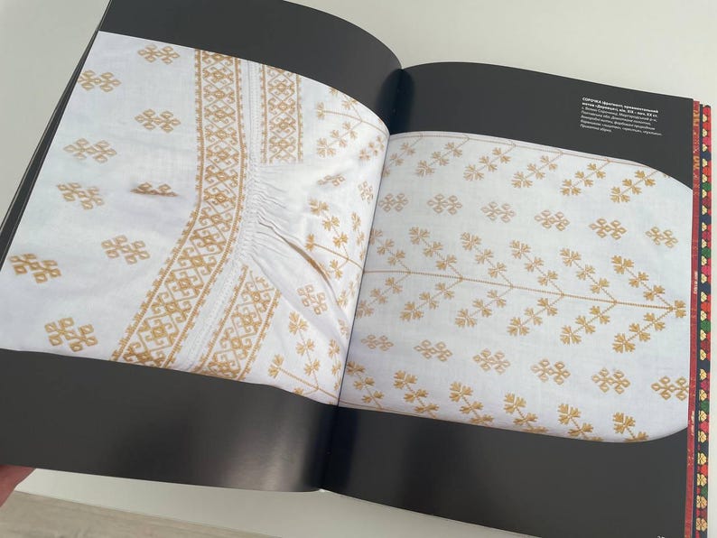 May include: Two pages from a book showing examples of white fabric with gold embroidery. The embroidery on the left page features a geometric pattern. The embroidery on the right page features a floral pattern.