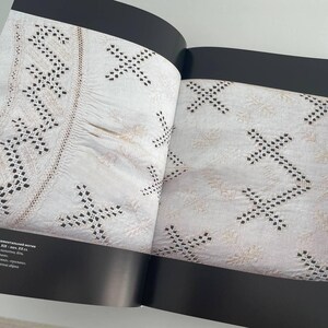 May include: A close-up of a book open to a page showing a white fabric with black embroidery. The fabric has a pattern of small crosses and a border of a more intricate design. The text on the page reads "СОРOЧKA (ручная вышивка, ornamented linen, 19th c., Ukrainian folk art, embroidered linen, 32 x 42 cm, private collection, Germany".