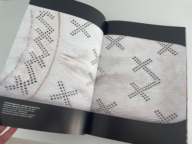 May include: A close-up of a book open to a page showing a white fabric with black embroidery. The fabric has a pattern of small crosses and a border of a more intricate design. The text on the page reads "СОРOЧKA (ручная вышивка, ornamented linen, 19th c., Ukrainian folk art, embroidered linen, 32 x 42 cm, private collection, Germany".