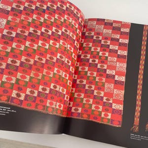 May include: A red and white checkered fabric with a geometric pattern. The fabric is woven with different colored threads, including red, white, green, and purple. The fabric is displayed on a book with the text "PLAHTA (blanket) - ethnic, ornamental, motif "Afghanskiy uzol" (Afghan knot), XIX - XX cc.  The fabric is made in the village of "Kishlak" (village) in the "Kishlak" (village) district of the "Kishlak" (village) region.  The fabric is made of wool and cotton.  The fabric is used for blankets, rugs, and other household items.  The fabric is a traditional Afghan textile.