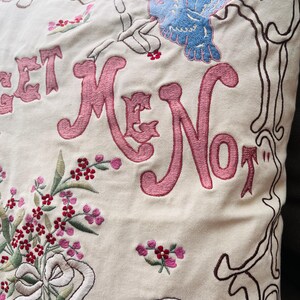 Op de afbeelding: Cr&egrave;mekleurig kussen met geborduurd bloemen- en vlinderontwerp. Het kussen heeft de woorden "Forget Me Not" in roze script. Een boeket bloemen en een lint zijn ook op het kussen geborduurd.