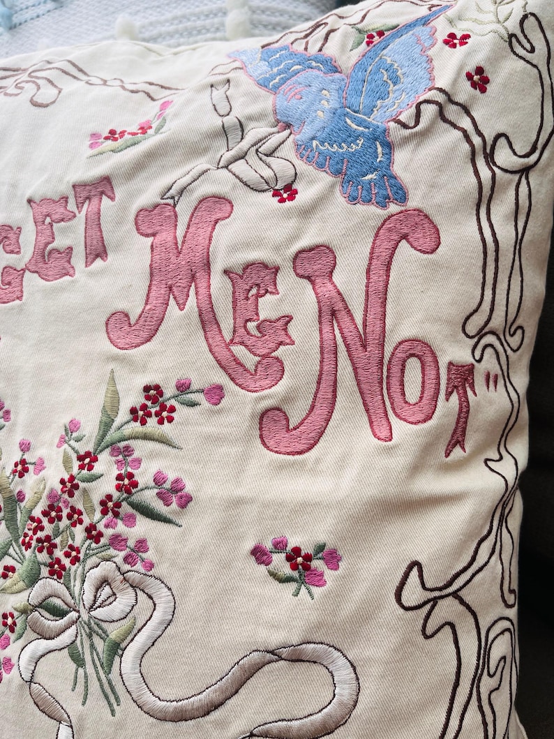 Op de afbeelding: Cr&egrave;mekleurig kussen met geborduurd bloemen- en vlinderontwerp. Het kussen heeft de woorden "Forget Me Not" in roze script. Een boeket bloemen en een lint zijn ook op het kussen geborduurd.