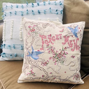 Op de afbeelding: Twee decoratieve kussens. E&eacute;n is wit met blauwe pompons en een geweven textuur. De andere is cr&egrave;mekleurig met geborduurde bloemen- en lintdetails, de woorden "Forget Me Not" en twee blauwe vogels.