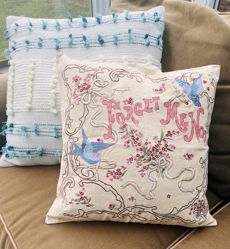 Op de afbeelding: Twee decoratieve kussens. E&eacute;n is wit met blauwe pompons en een geweven textuur. De andere is cr&egrave;mekleurig met geborduurde bloemen- en lintdetails, de woorden "Forget Me Not" en twee blauwe vogels.