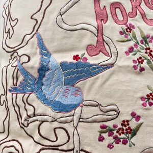 Op de afbeelding: Detail van geborduurde stof met een blauwe vogel en roze bloemmotieven. Het woord "FORGE" is geborduurd in roze letters. Het ontwerp is omlijnd met bruin en wit garen op een gebroken witte achtergrond. Metrische maten zijn niet van toepassing.