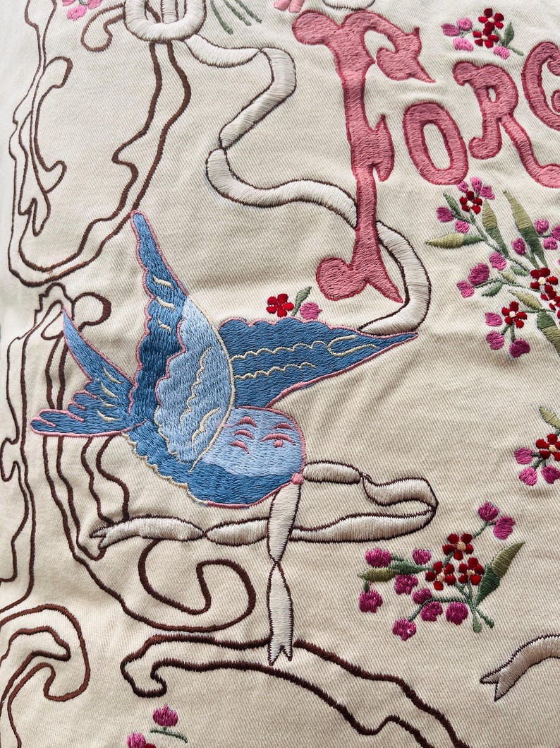Op de afbeelding: Detail van geborduurde stof met een blauwe vogel en roze bloemmotieven. Het woord "FORGE" is geborduurd in roze letters. Het ontwerp is omlijnd met bruin en wit garen op een gebroken witte achtergrond. Metrische maten zijn niet van toepassing.