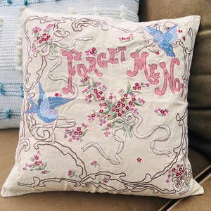 Op de afbeelding: Cr&egrave;mekleurig sierkussen met geborduurd bloemen- en vogelontwerp. De woorden "Forget Me Not" zijn roze, met blauwe vogels en roze bloemen. De borduursels zijn bruin omlijnd. Decoratief woonaccessoire.