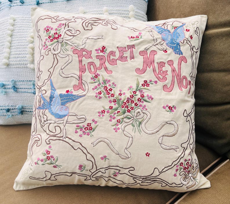 Op de afbeelding: Cr&egrave;mekleurig sierkussen met geborduurd bloemen- en vogelontwerp. De woorden "Forget Me Not" zijn roze, met blauwe vogels en roze bloemen. De borduursels zijn bruin omlijnd. Decoratief woonaccessoire.