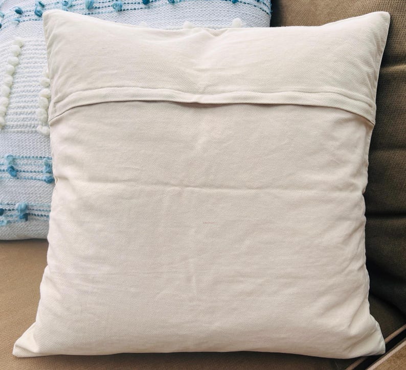 Op de afbeelding: Een vierkant, cr&egrave;mekleurig kussen met een getextureerd oppervlak. Het kussen is gemaakt van een zachte stof en heeft een eenvoudig, minimalistisch ontwerp. Het kussen maakt deel uit van een woondecoratiecollectie.