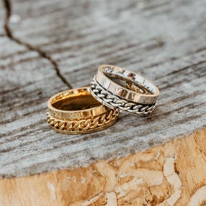 May include: Two gold and silver spinner rings with chain details. The rings are stacked on top of each other on a wooden surface.