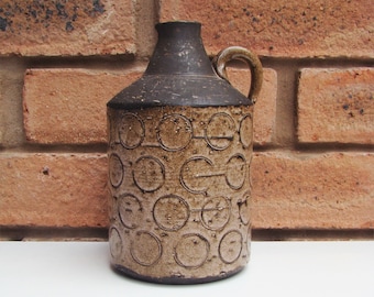 May include: A brown and beige ceramic vase with a circular pattern. The vase has a handle and a narrow neck.