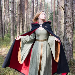 May include: A woman wearing a long green dress with a black and red hooded cloak. The cloak has a gold buckle and a brown lining. She is standing in a forest.