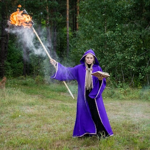 May include: A person in a purple hooded robe with gold trim holds a flaming torch and a book in a forest setting. The robe has long sleeves and a gold rope belt. The torch emits smoke and bright flames.