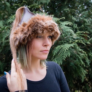 Può includere: Un cappello di pelliccia sintetica marrone con una parte superiore in pelle grigia e un lungo portacoda di cavallo. Il cappello è indossato da una persona con i capelli verdi e biondi.