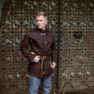 Pode incluir: Uma jaqueta acolchoada marrom com fivelas pretas e um cinto de couro marrom. A jaqueta é combinada com calças cinzas e botas de couro marrom. O fundo apresenta uma grande porta de metal ornamentada.