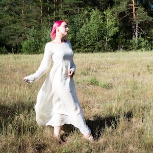 May include: A woman with pink hair wearing a white linen dress walks through a field of tall grass.