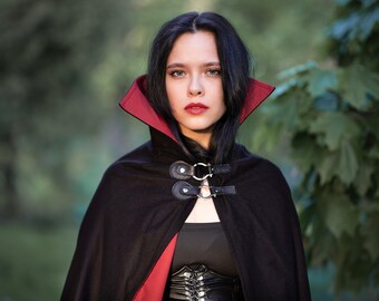 May include: A woman wearing a black cape with a red lining and a silver buckle. The cape has a high collar. She is looking directly at the camera.