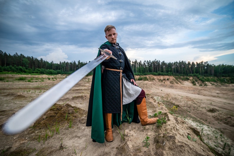 Peut inclure: Un homme v&ecirc;tu d'un costume m&eacute;di&eacute;val vert et noir tient une &eacute;p&eacute;e devant lui. Il se tient sur une colline sablonneuse avec une for&ecirc;t en arri&egrave;re-plan.