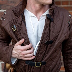 May include: A brown quilted jacket with diamond stitching, featuring black leather straps with gold buckles. The jacket has a high collar and is worn over a white shirt. The background is a brick wall.
