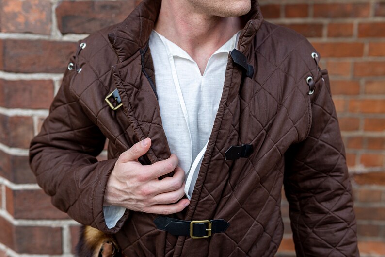 May include: A brown quilted jacket with diamond stitching, featuring black leather straps with gold buckles. The jacket has a high collar and is worn over a white shirt. The background is a brick wall.