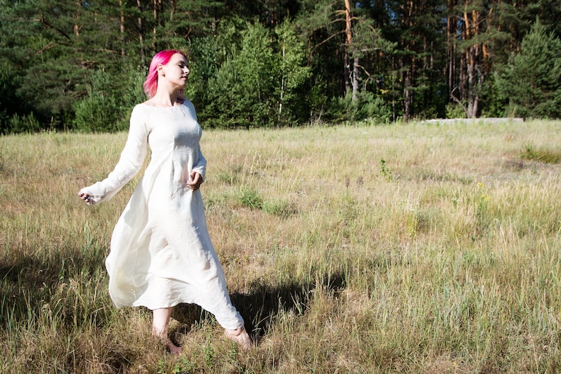 May include: A woman with pink hair wearing a white linen dress walks through a field of tall grass.