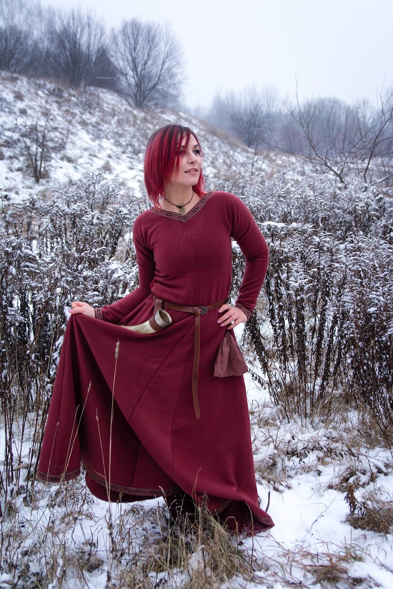 May include: A woman with red hair wearing a long, burgundy dress with a brown belt and a brown pouch. She is standing in a snowy field with trees in the background.