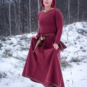 May include: A woman with red hair wearing a burgundy medieval dress with a brown belt and a horn drinking vessel. She is standing in a snowy forest.