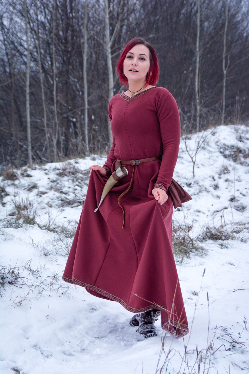 May include: A woman with red hair wearing a burgundy medieval dress with a brown belt and a horn drinking vessel. She is standing in a snowy forest.