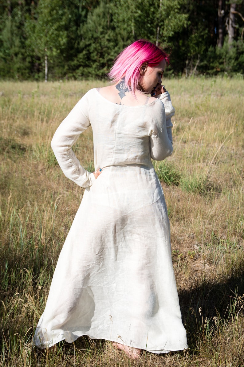 May include: A woman with pink hair wearing a white linen dress stands in a field of tall grass.