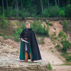 Peut inclure: Un homme v&ecirc;tu d'une cape &agrave; capuche noire et verte se tient sur une colline sablonneuse avec une &eacute;p&eacute;e &agrave; la main. Il regarde de c&ocirc;t&eacute;.