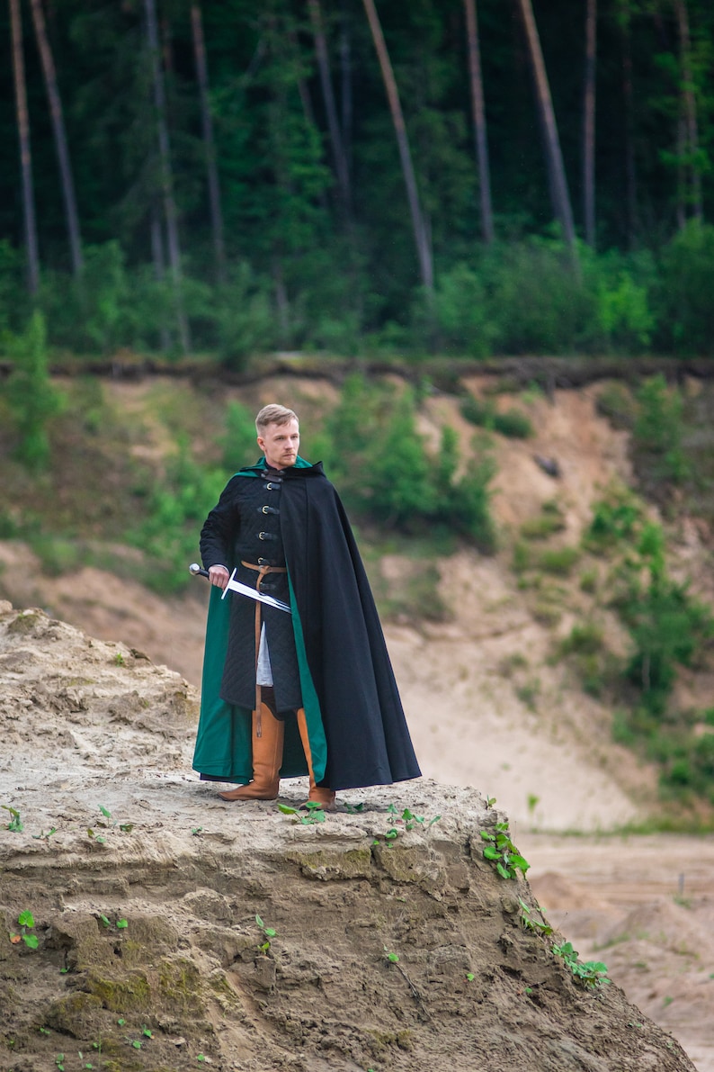 Peut inclure: Un homme v&ecirc;tu d'une cape &agrave; capuche noire et verte se tient sur une colline sablonneuse avec une &eacute;p&eacute;e &agrave; la main. Il regarde de c&ocirc;t&eacute;.