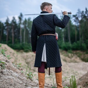 Peut inclure: Un homme portant un manteau matelass&eacute; noir avec une ceinture marron et des bottes en cuir marron, tenant une &eacute;p&eacute;e sur son &eacute;paule.