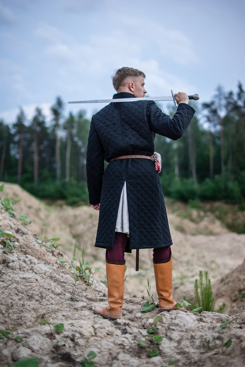 Peut inclure: Un homme portant un manteau matelass&eacute; noir avec une ceinture marron et des bottes en cuir marron, tenant une &eacute;p&eacute;e sur son &eacute;paule.
