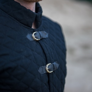 Peut inclure: Une veste matelass&eacute;e noire avec un col montant et deux boucles en laiton. La veste est faite d'un tissu &eacute;pais et r&eacute;sistant et a un design classique.