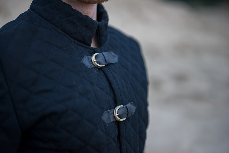 Peut inclure: Une veste matelass&eacute;e noire avec un col montant et deux boucles en laiton. La veste est faite d'un tissu &eacute;pais et r&eacute;sistant et a un design classique.