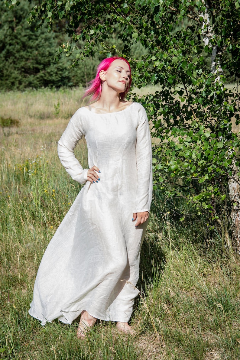 May include: A woman with pink hair wearing a white linen dress stands in a field of green grass. The dress has long sleeves and a flowing skirt.