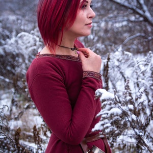 May include: A woman with bright red hair wearing a burgundy dress with a brown belt and a horn drinking vessel. She is standing in a snowy forest.