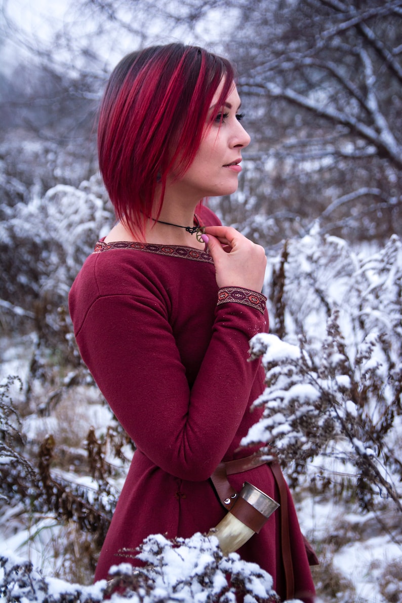 May include: A woman with bright red hair wearing a burgundy dress with a brown belt and a horn drinking vessel. She is standing in a snowy forest.