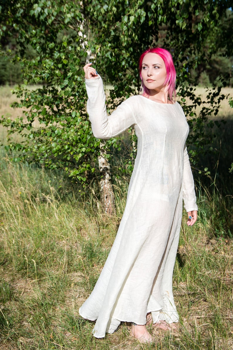 May include: A woman with pink hair wearing a long, white linen dress stands in a field of tall grass. The dress has long sleeves and a simple, loose fit.
