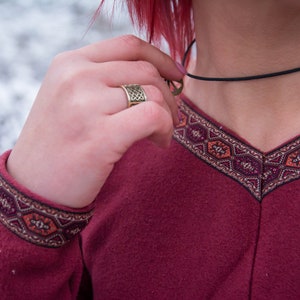 May include: A burgundy long-sleeved shirt with a geometric pattern around the neckline and cuffs. The shirt has a v-neck and a black choker necklace.