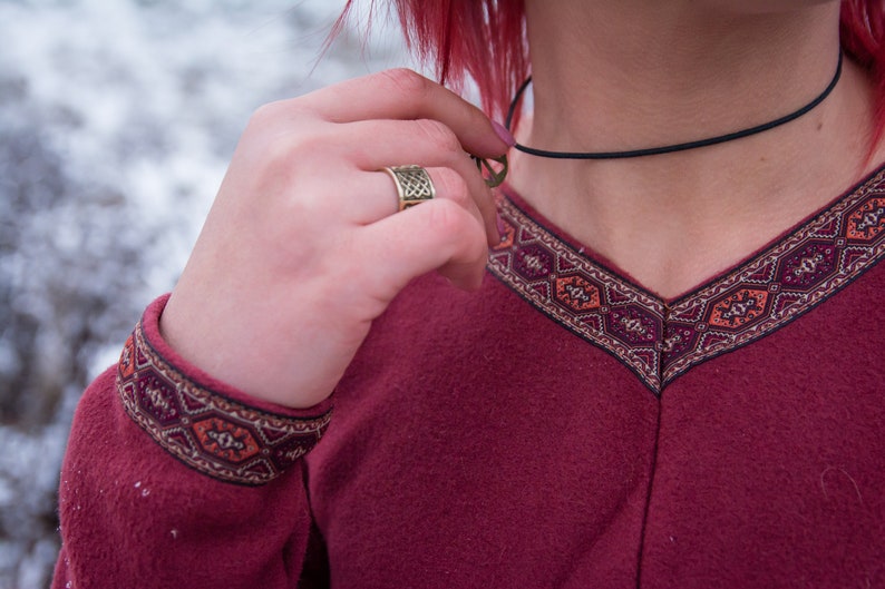 May include: A burgundy long-sleeved shirt with a geometric pattern around the neckline and cuffs. The shirt has a v-neck and a black choker necklace.