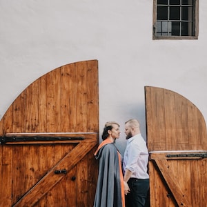 May include: A couple stands facing each other in front of two wooden doors. The woman is wearing a gray hooded cape with an orange lining. The man is wearing a white button-down shirt and black pants.