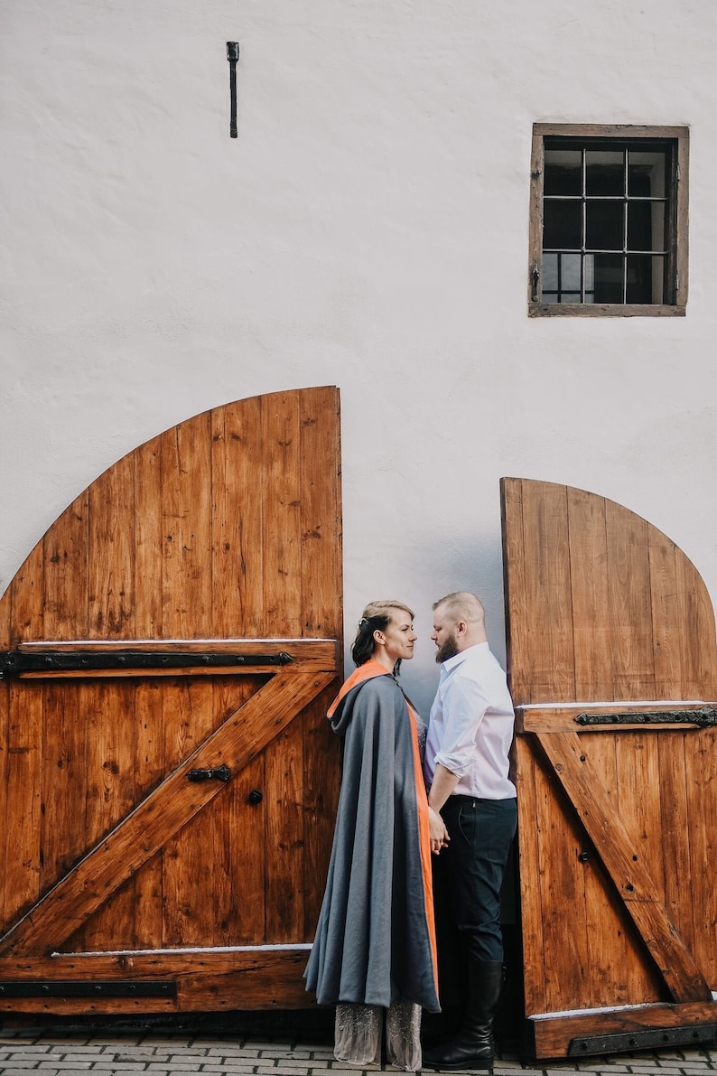 May include: A couple stands facing each other in front of two wooden doors. The woman is wearing a gray hooded cape with an orange lining. The man is wearing a white button-down shirt and black pants.