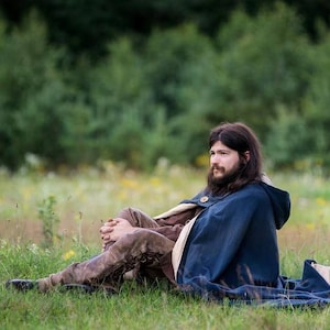 May include: A man with long brown hair and a beard is sitting in a field wearing a blue hooded cloak and brown pants. He is looking off to the side.