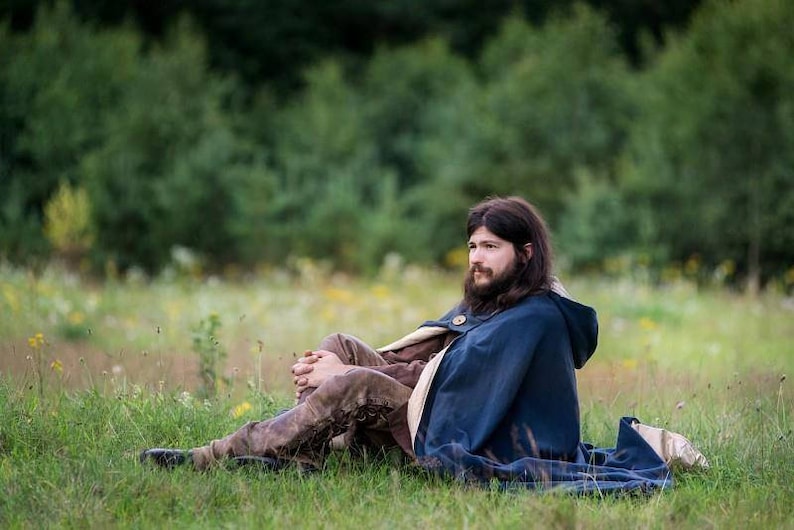 May include: A man with long brown hair and a beard is sitting in a field wearing a blue hooded cloak and brown pants. He is looking off to the side.