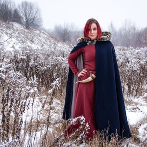 May include: A woman with red hair wearing a long, dark blue cloak with a fur-trimmed hood. She is standing in a snowy field with tall, brown grasses.