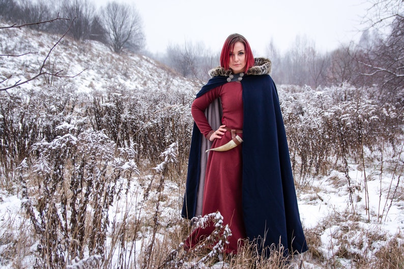 May include: A woman with red hair wearing a long, dark blue cloak with a fur-trimmed hood. She is standing in a snowy field with tall, brown grasses.
